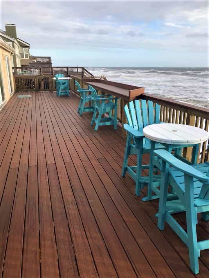 Wide stained wood deck with turquoise chairs and a rail-top bar overlooking breaking ocean surf