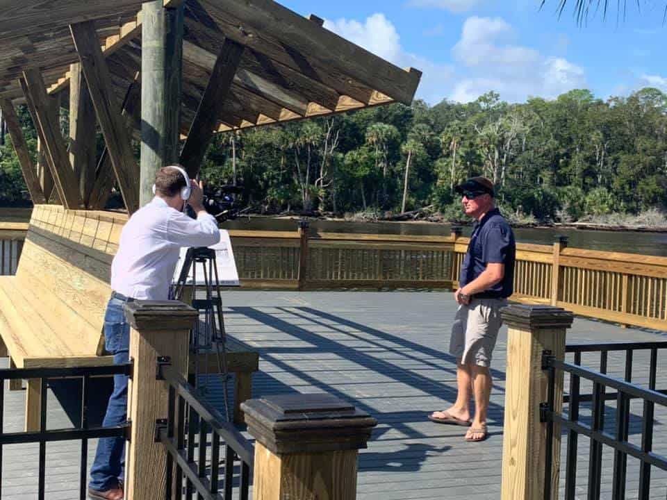 Man interviewed on camera on a wharf deck beside a timber pavilion and wood railings