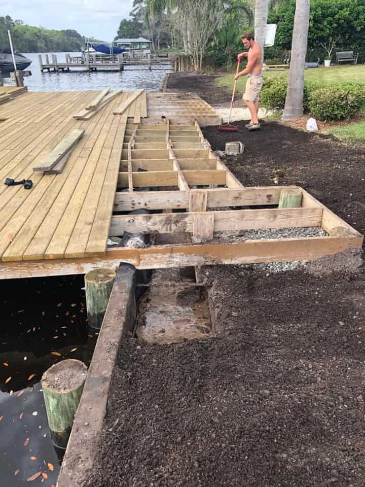 Man rakes soil beside an open deck frame of joists next to a waterfront dock