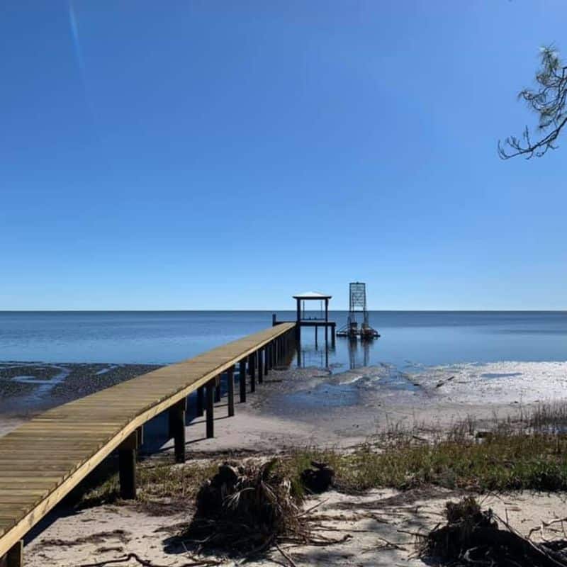 Wooden walkway crosses sand to a covered dock and boat lift standing in calm water