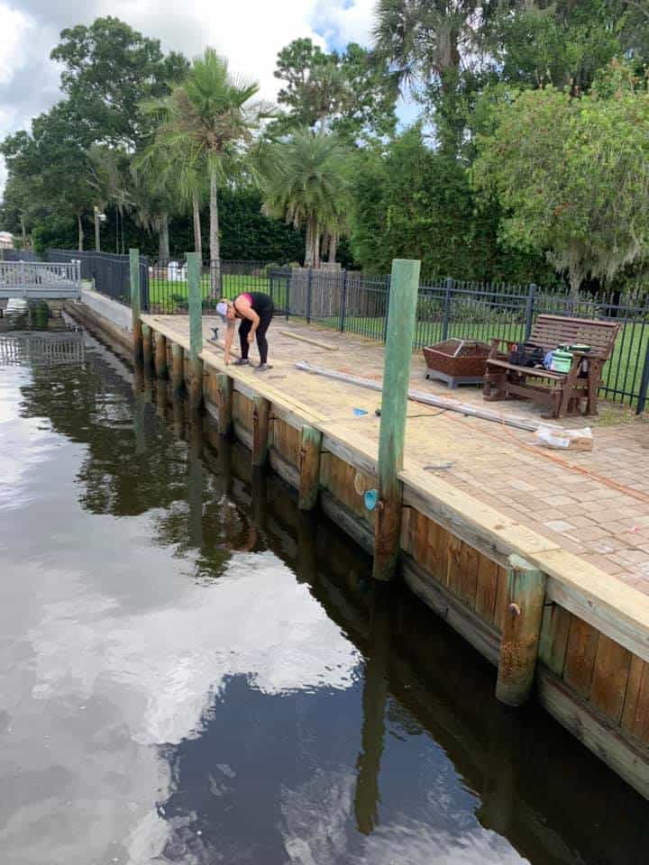 Person working on a timber seawall cap beside a paver patio and dark canal water
